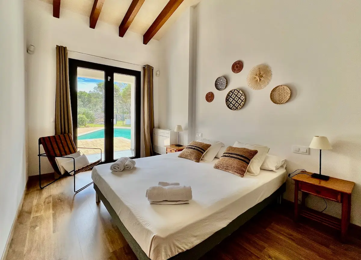 Bright Mediterranean-style bedroom in a Cala Morell villa featuring white walls, exposed wooden ceiling beams, and large glass doors opening directly to a private swimming pool.