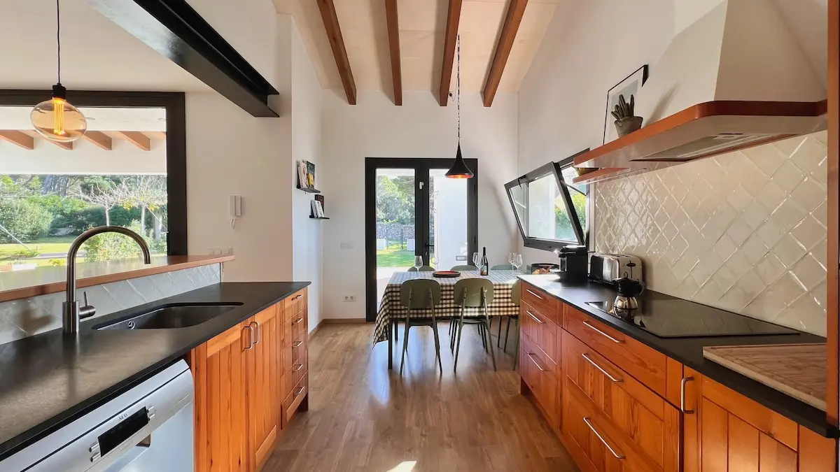 Modern open-plan kitchen with wooden cabinetry, black countertops, and a dining table set for guests, leading to a sunlit patio in a Menorca vacation home.