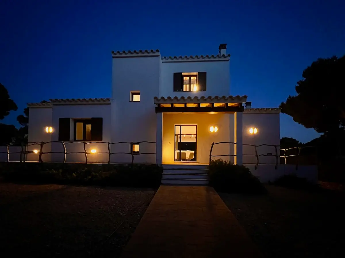 Exterior view of a traditional white-washed Menorcan villa at night, illuminated by warm lights with a stone path and rustic wooden fencing.