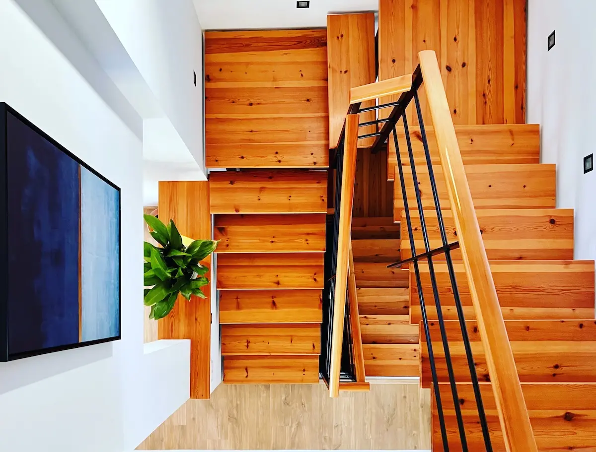 op-down view of a modern architectural wooden staircase with black metal railings, featuring a large blue abstract painting and indoor greenery in a luxury Menorcan home