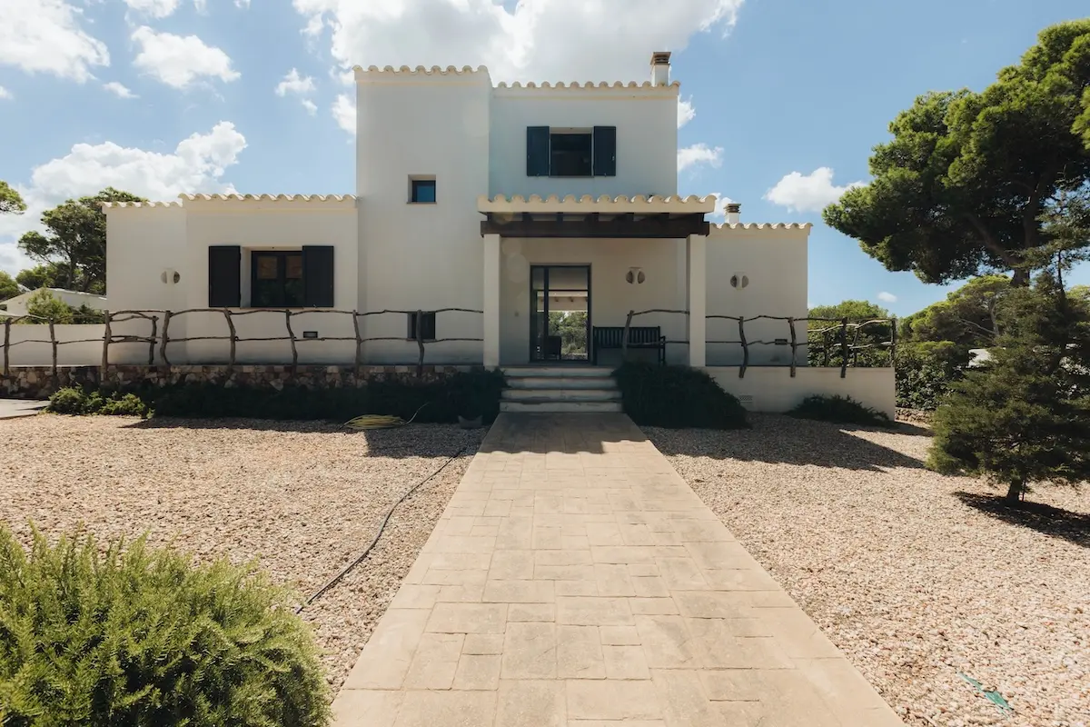 Daytime view of a traditional white-washed villa in Cala Morell, Menorca, with a paved walkway, blue shutters, and surrounding pine trees under a clear blue sky