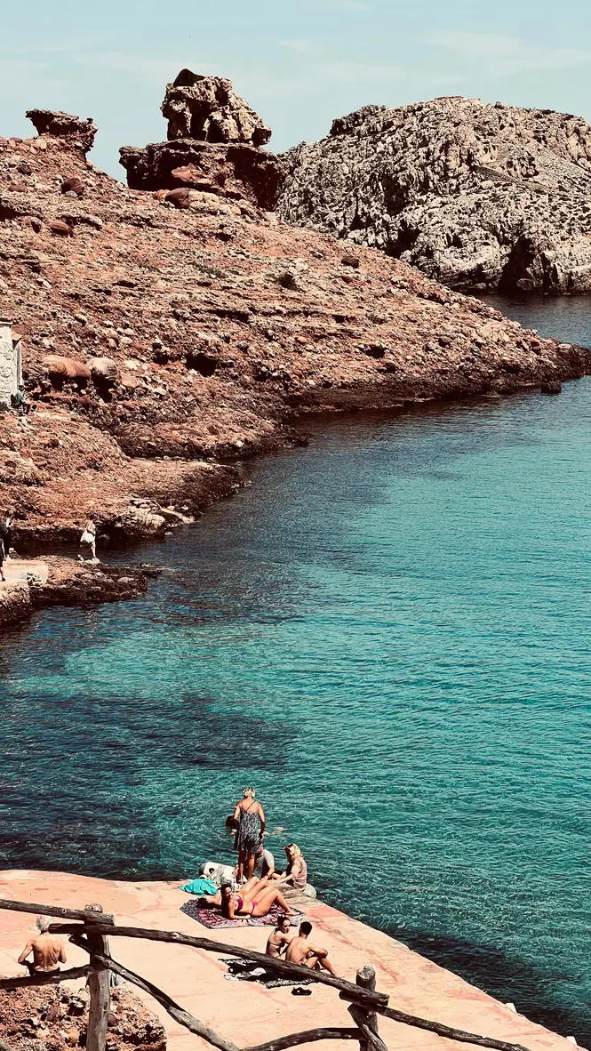 Travelers sunbathing on the rocky platforms of Cala Morell beach, surrounded by turquoise Mediterranean waters and dramatic red-toned cliffs