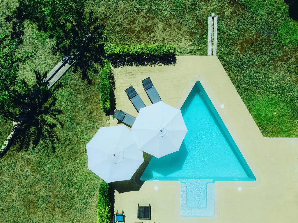 Aerial drone shot of a unique triangular-shaped swimming pool with turquoise water, white umbrellas, and lounge chairs surrounded by a lush green lawn
