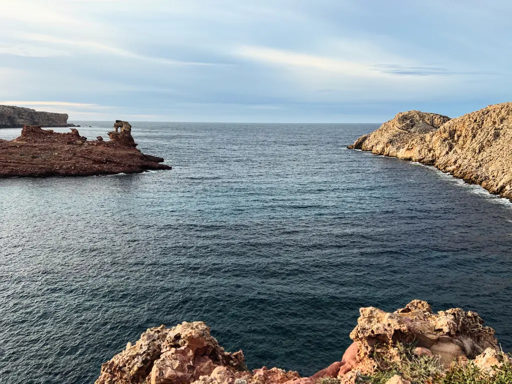 wide scenic view of the dramatic red-toned rocky cliffs and deep blue Mediterranean Sea at the entrance of Cala Morell bay in Menorca, Spain.