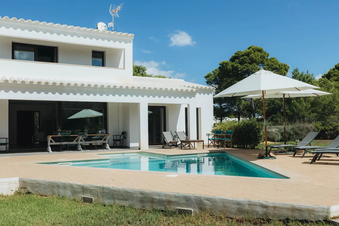 Terrace and pool area at Villa Arrow in Menorca, featuring white parasols, sun loungers, and an outdoor dining table under the shaded porch.