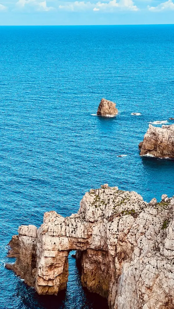 A vertical shot of dramatic limestone cliffs and a natural rock arch formation over the vibrant turquoise Mediterranean Sea in Cala Morell, Menorca.