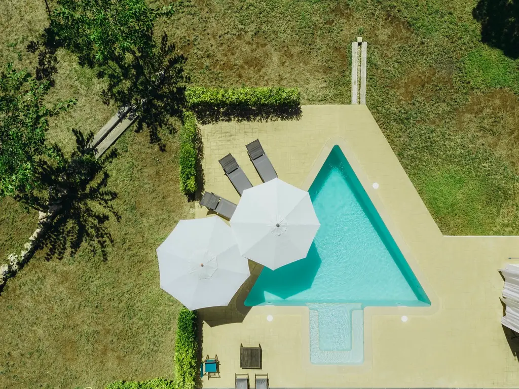 Top-down aerial view of the signature arrow-shaped swimming pool at Villa Arrow, Menorca, with white parasols and lounge chairs surrounded by lawn.