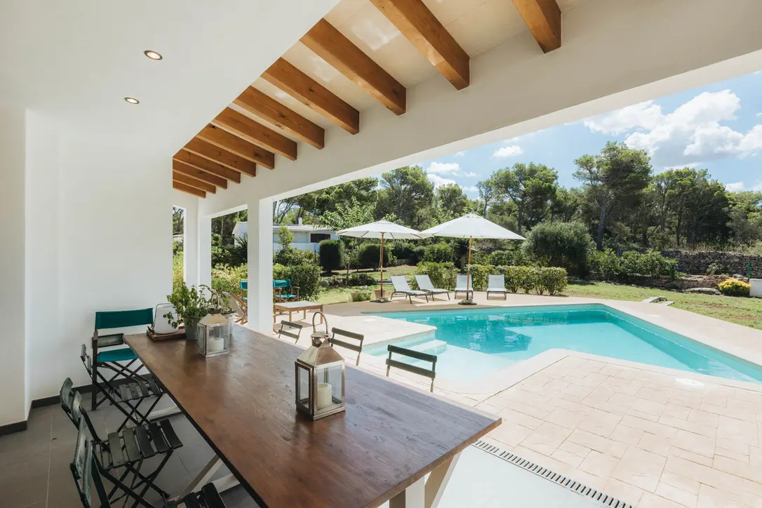 Shaded outdoor dining area at Villa Arrow featuring a long wooden table, exposed timber beams, and a view of the turquoise swimming pool and Mediterranean garden.