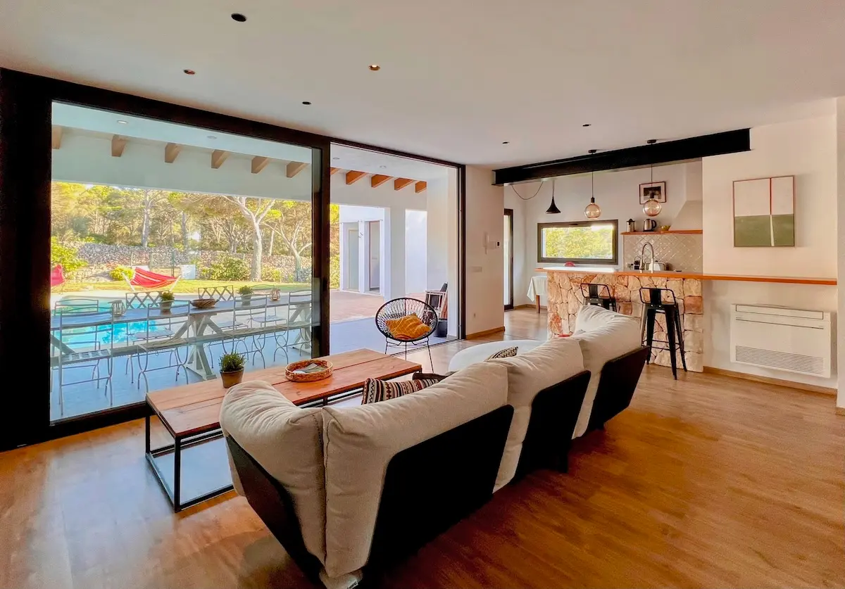 Modern open-plan interior of Villa Arrow showing a comfortable living room with a white sofa, a stone-accented breakfast bar, and large glass sliding doors opening to the pool area.